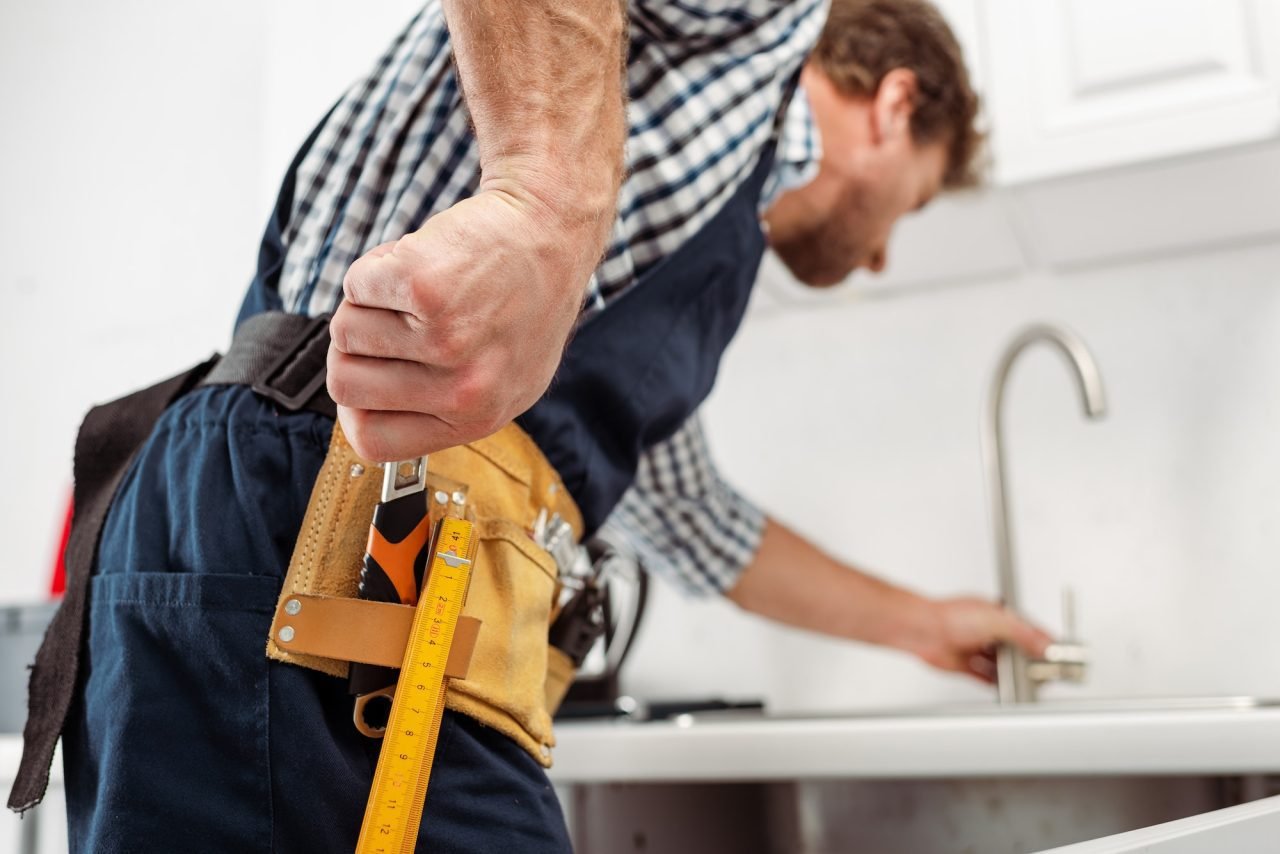 selective-focus-of-plumber-taking-wrench-from-tool-belt-while-fixing-faucet-in-kitchen